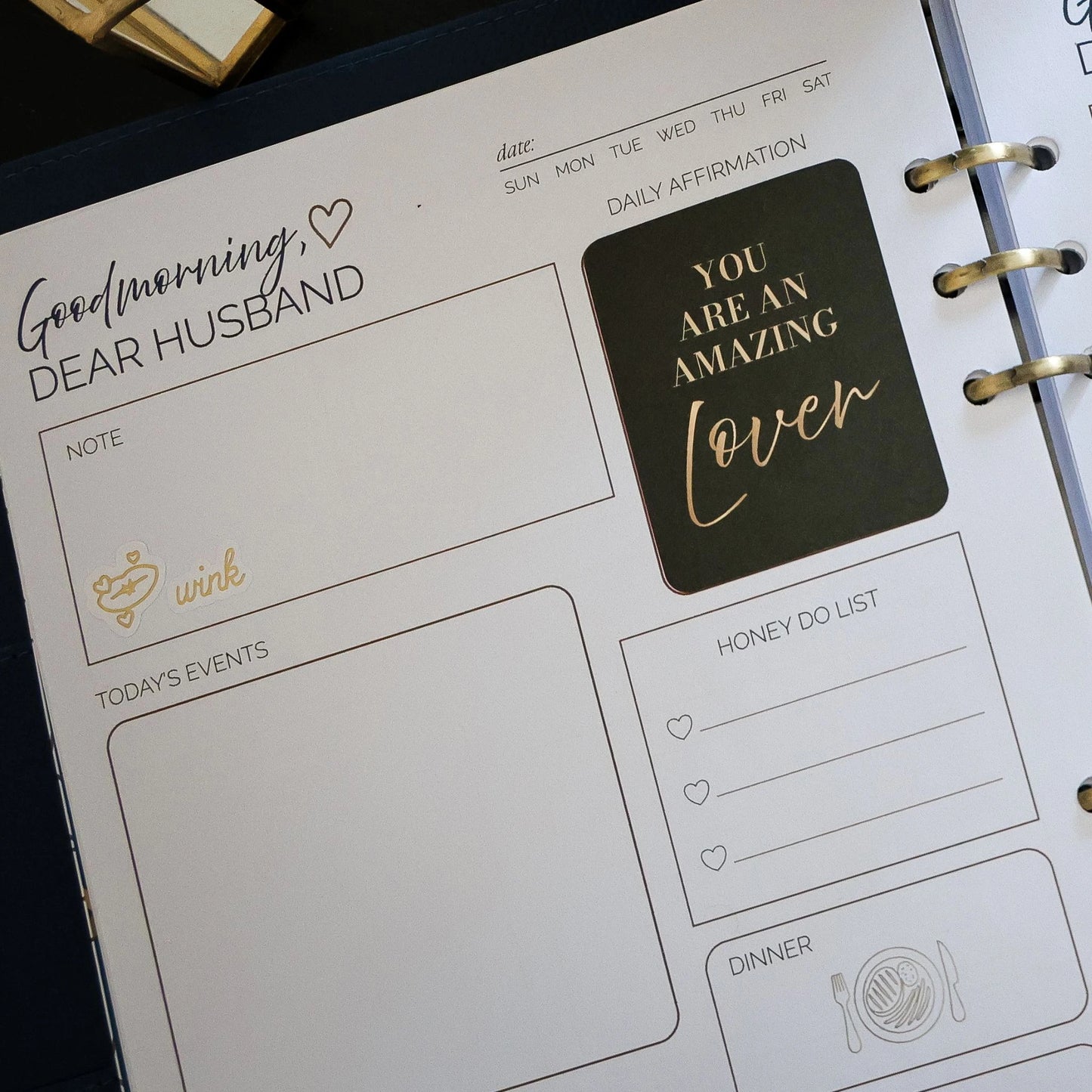 A close-up of a Honor Him Thru Intimacy Affirmation Sticker Book page with a 'Good morning, Dear Husband' layout. The page includes fields for notes, today's events, a honey do list, dinner, a daily affirmation sticker that reads 'You are an amazing lover' in gold on black, and a gold 'wink' doodle.